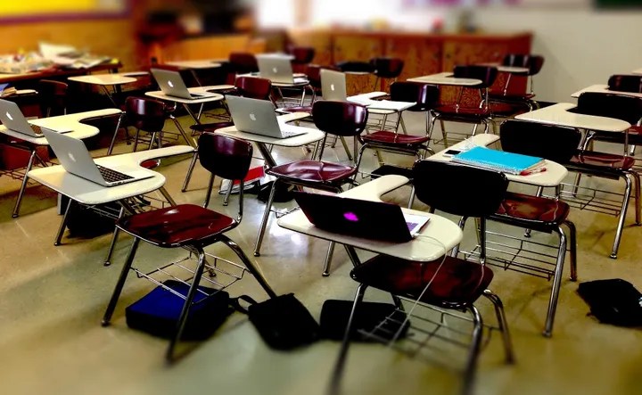 Photograph of a classroom with individual desks. Many desks have open laptops on them.